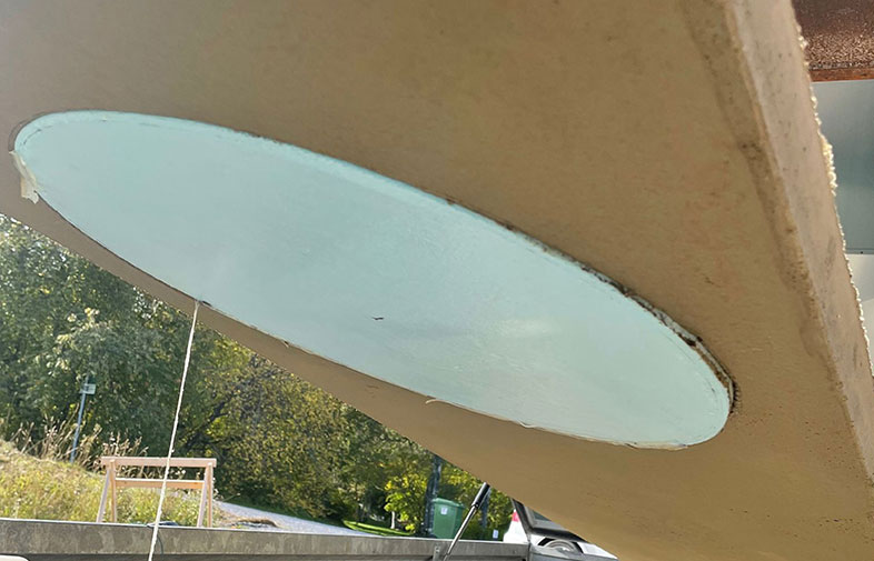 Beige planka i sned vinkel med ljusblå oval på undersidan, lövträd i bakgrunden.