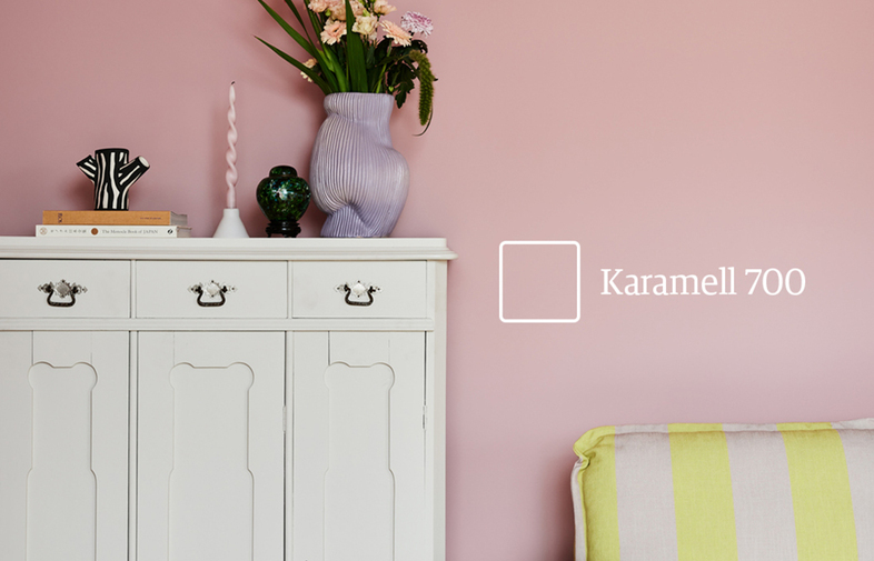 Pastellrosa vägg med färgkod Karamell 700, vitt skåp, lila vas med blommor, gulrandig soffa.