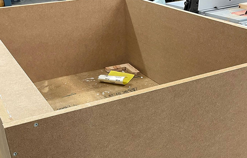 Öppen brun MDF-låda i verkstad med gul förpackning och träbitar inuti.