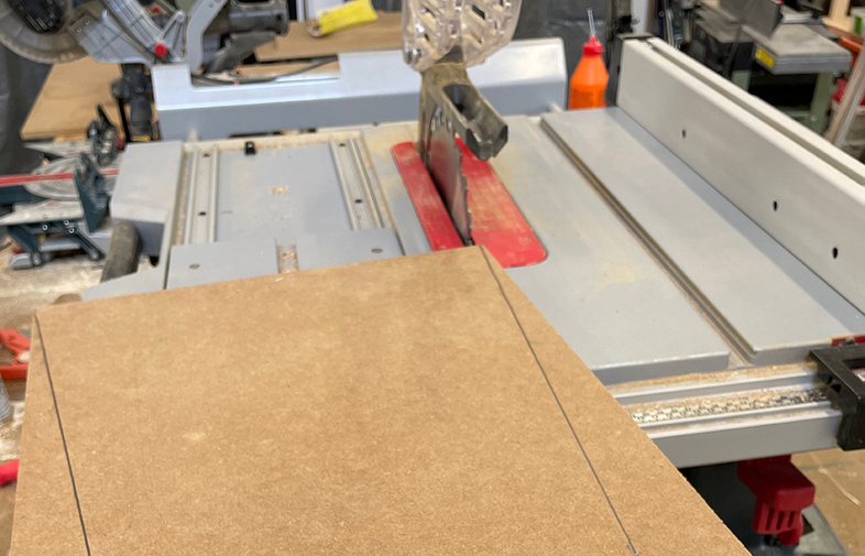 Grå bordsåg med röd insats, sågklinga synlig, spånskiva framför, orange limflaska i bakgrunden.