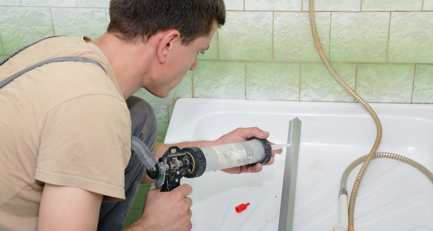 Hantverkare i beige tröja tätar vit badkarskant med fogpistol mot grönmönstrade kakelplattor.