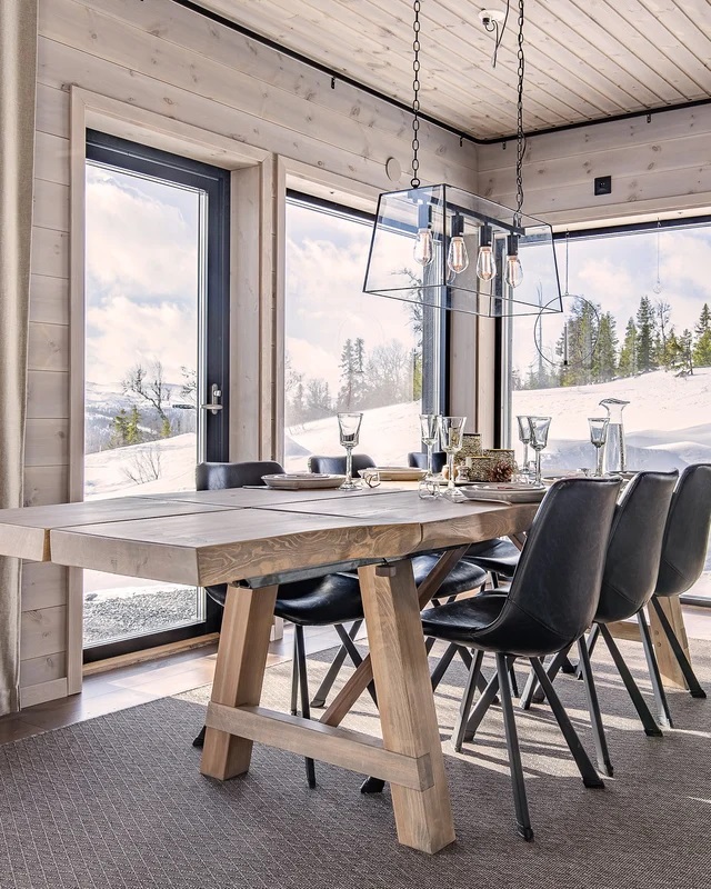 Ljust träbord med svarta stolar och hängande glaskupa framför stora fönster mot snölandskap.