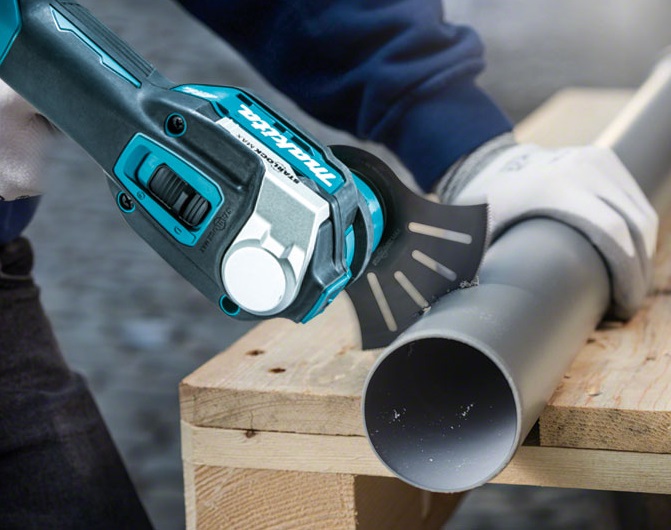 Turkos-svart oscillerande multiverktyg kapar grå plastledning på träbänk, handskbeklädd