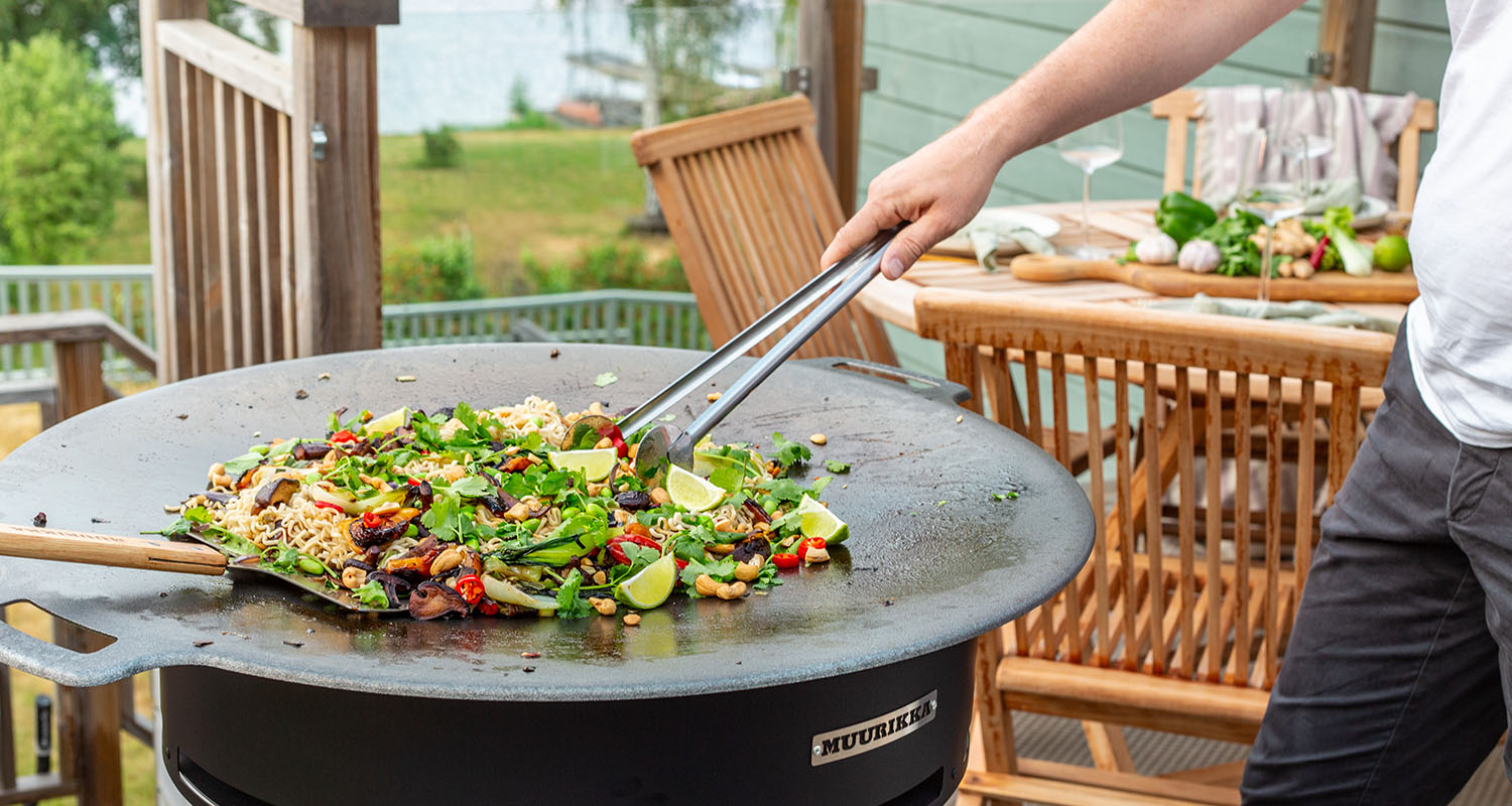Hand med tång rör färgglad grönsaks- och nudelwok med lime på rund utegrill på träaltan vid sjö.