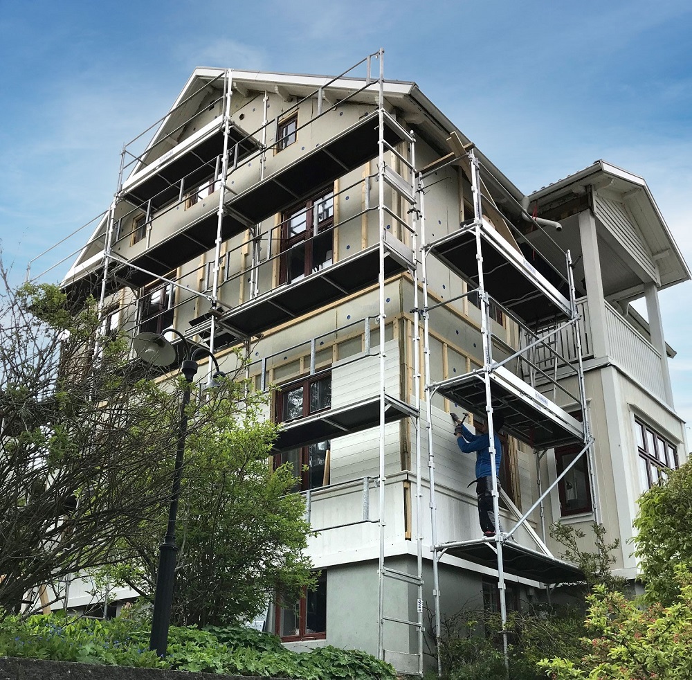 Vit trävilla under renovering med byggnadsställning, blå himmel, gröna buskar och arbetare.