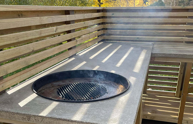 Betongbord med rund grillgrop, rökstigande gallergaller och trästaket i soligt höstljus.