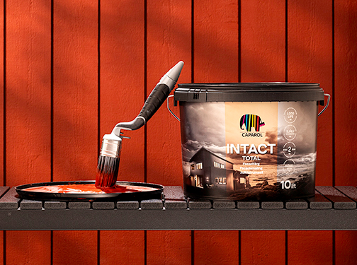 Svart målarhink med färgglad Caparol-logga och pensel doppad i röd färg mot röd träpanel.