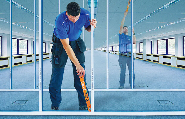 Man i blå tröja mäter glasvägg i tomt, ljust kontorslandskap.