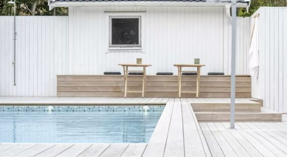 Vit villa med pool, ljust trädäck, två små träbord, stolar och gröna muggar.