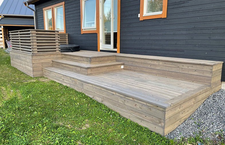 Trädäck i ljusgrått trä med flera nivåer framför svart panelhus med orange fönsterramar.