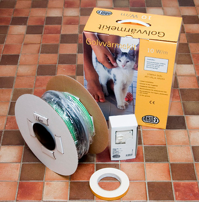 Gul golvvärmebox med kattbild, grön kabelrulle, vit termostat och tejprulle på klinkergolv.