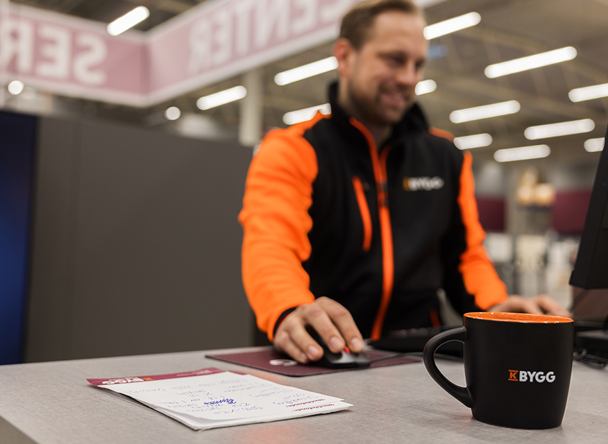Medarbetare på K-Bygg arbetar vid kassan med dator och mus. På disken ligger anteckningar och en kaffemugg med K-Byggs logotyp, i en modern butiksmiljö.