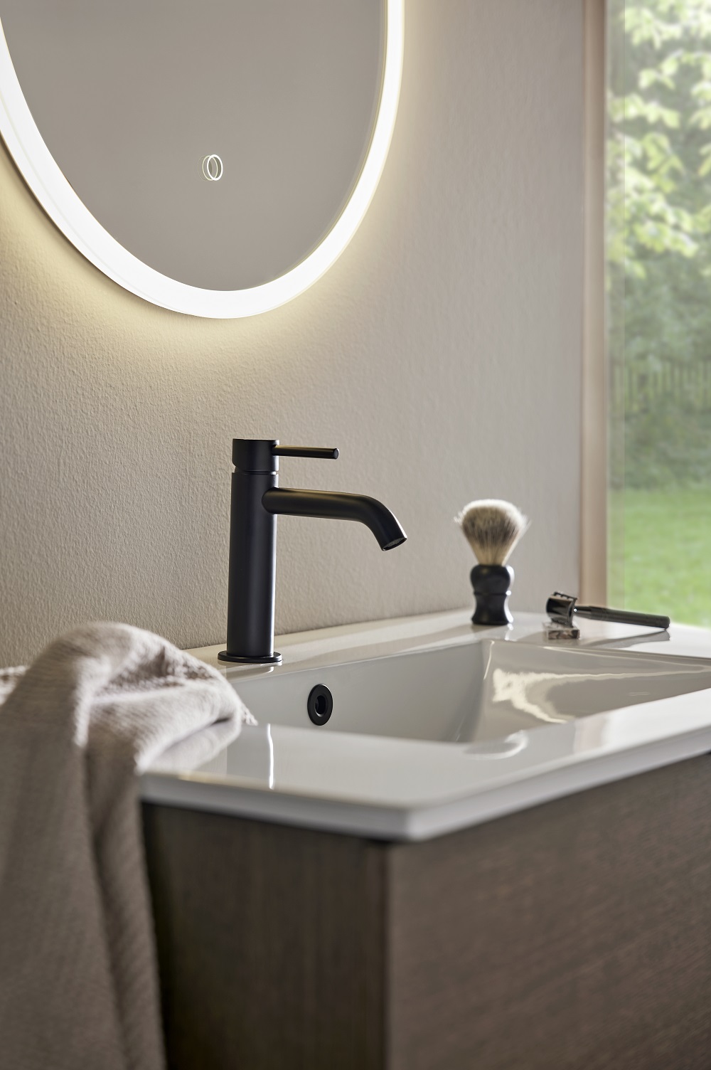 Rund LED-spegel över vitt handfat med svart kran, rakborste, rakhyvel och grå handduk.