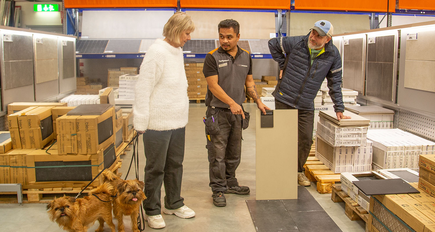Kund med två små bruna hundar och anställd i byggvaruhus tittar på gråbeige kakelplatta.