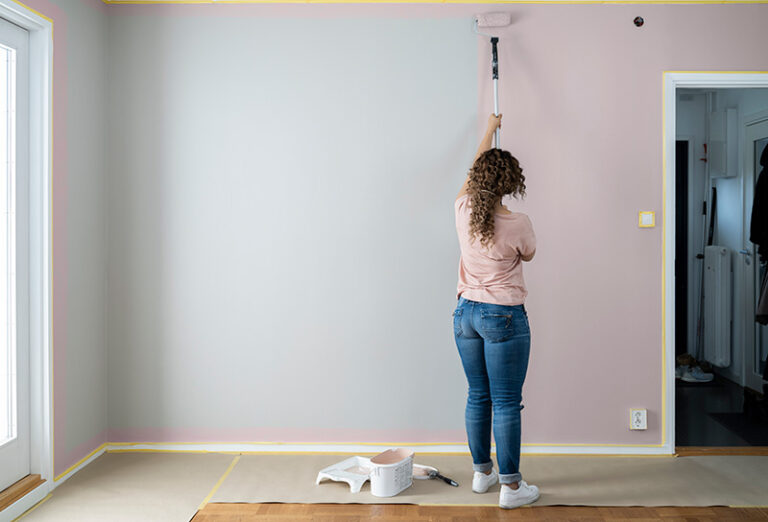 Kvinna målar inomhusvägg rosa över grå grund med roller på förlängningsskaft, golv skyddat.