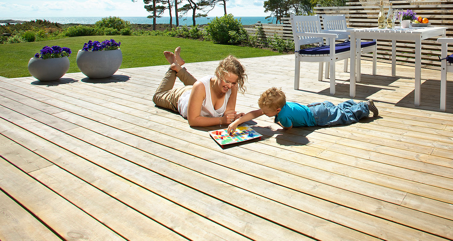 Kvinna och barn ligger på ljust trädäck, spelar färgglatt brädspel med havsutsikt.