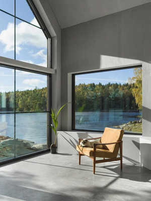 Ljust modernt rum med stora fönster mot sjö och skog, träfåtölj i cognacsbrunt läder.