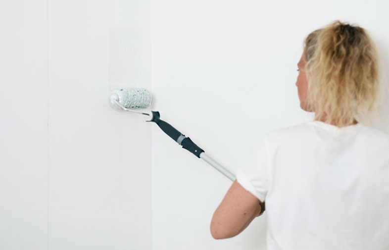 Kvinna med blont hår målar en vit vägg med roller på långt skaft.