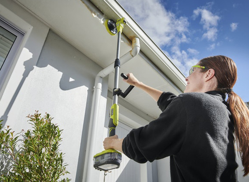 Person med långt hår använder grön batteridriven borste för rengöring under husets takfot.