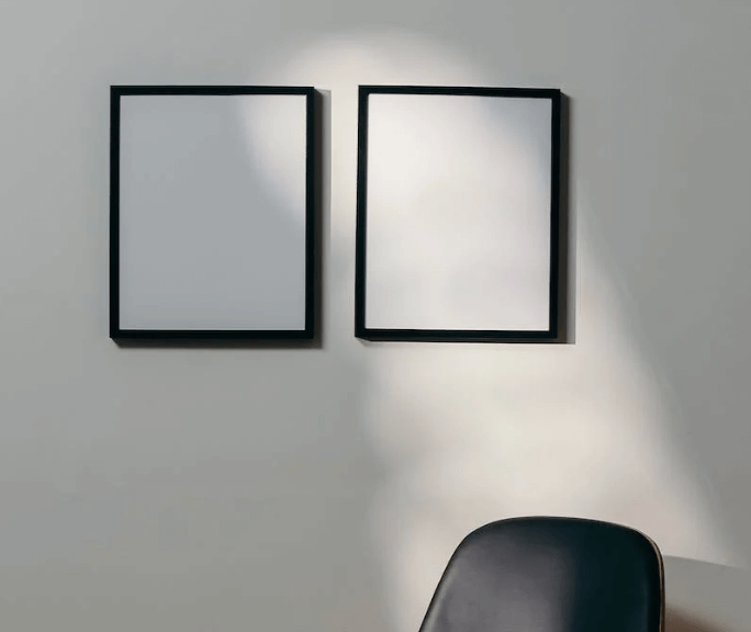 Två svarta tomma ramar på vit vägg i strålkastarljus, svart stol i hörnet.