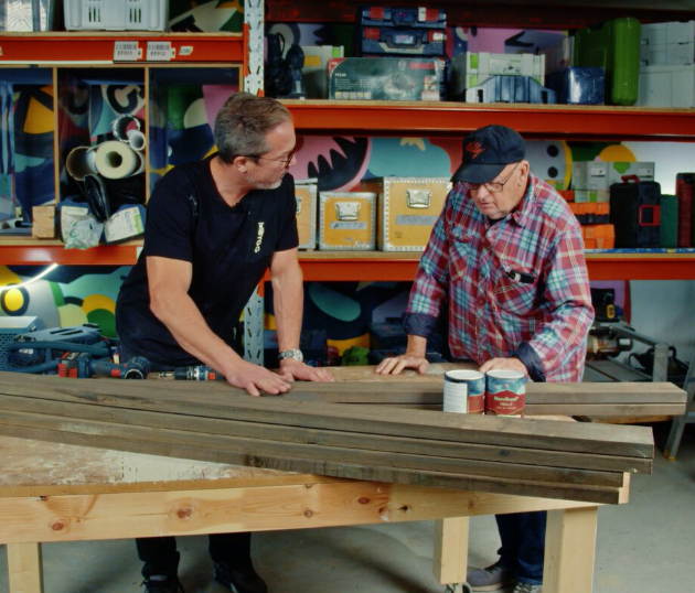 Två män lutar sig över träplankor på arbetsbänk i färgglad verkstad med verktyg och färgburkar