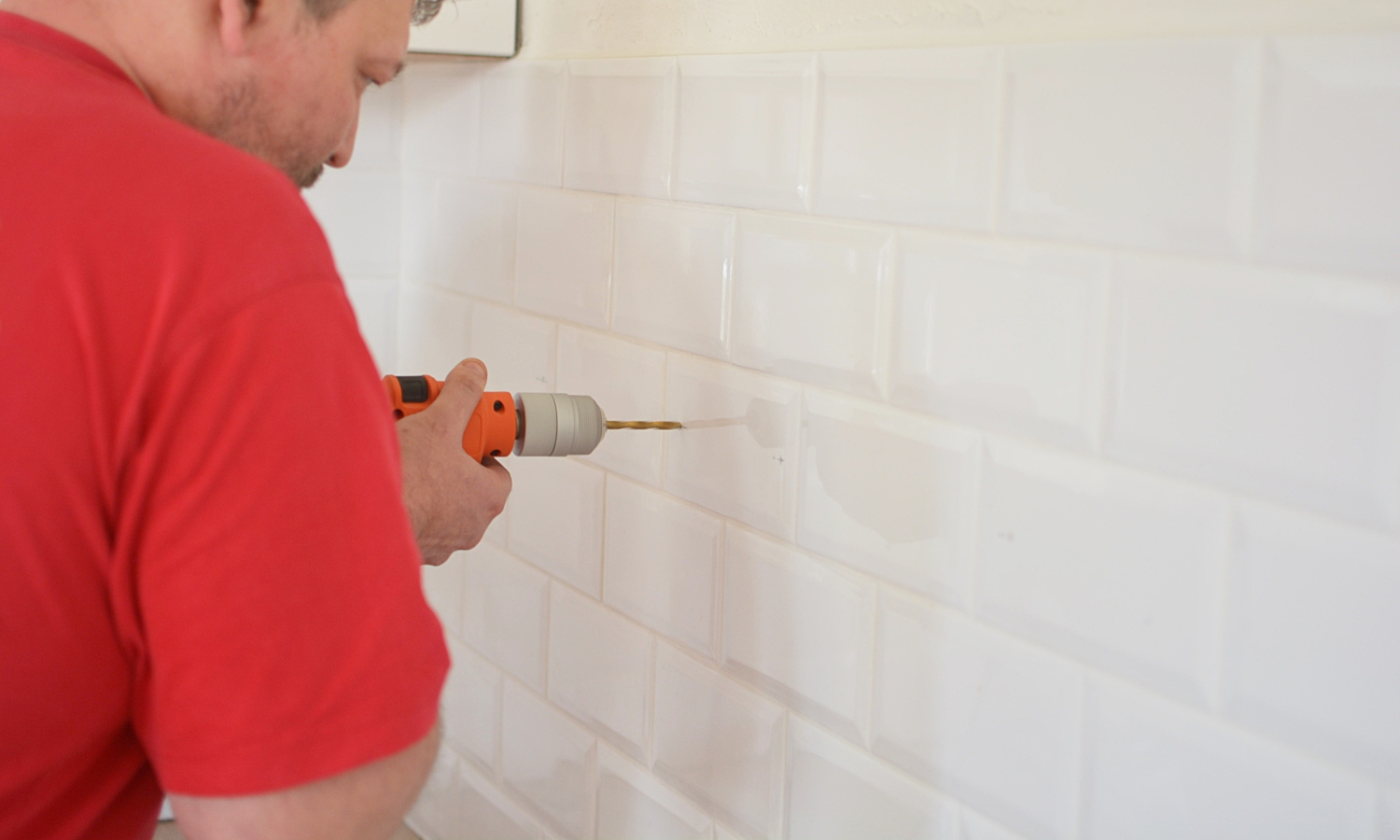 Person i röd t-shirt borrar med orange borrmaskin i vit kakelvägg