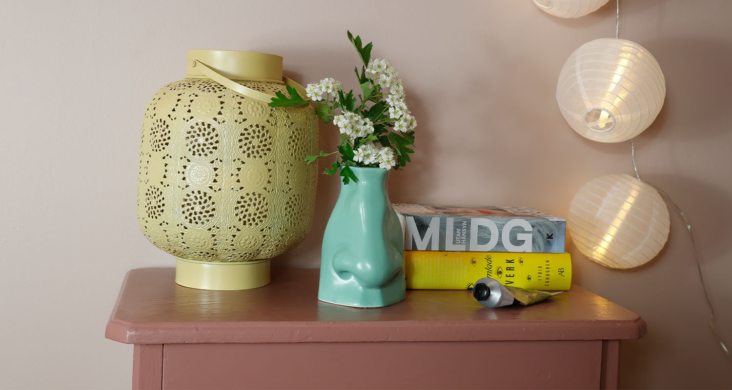 Rosa bord med gul perforerad lykta, grön näsvas med vita blommor, böcker och ljusslinga