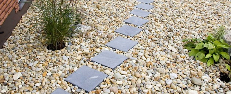 Grå skifferplattor som gång över beige småsten, med prydnadsgräs och hostor vid sidan.