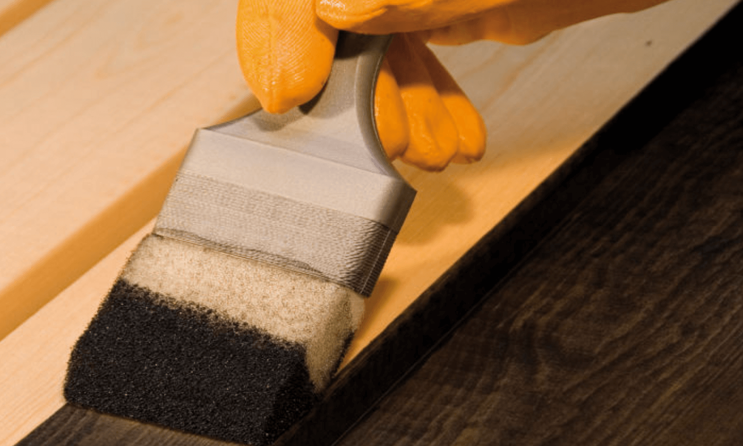 Orangehandskad hand betsar mörkt trä med skumapplikator på ljus träskiva
