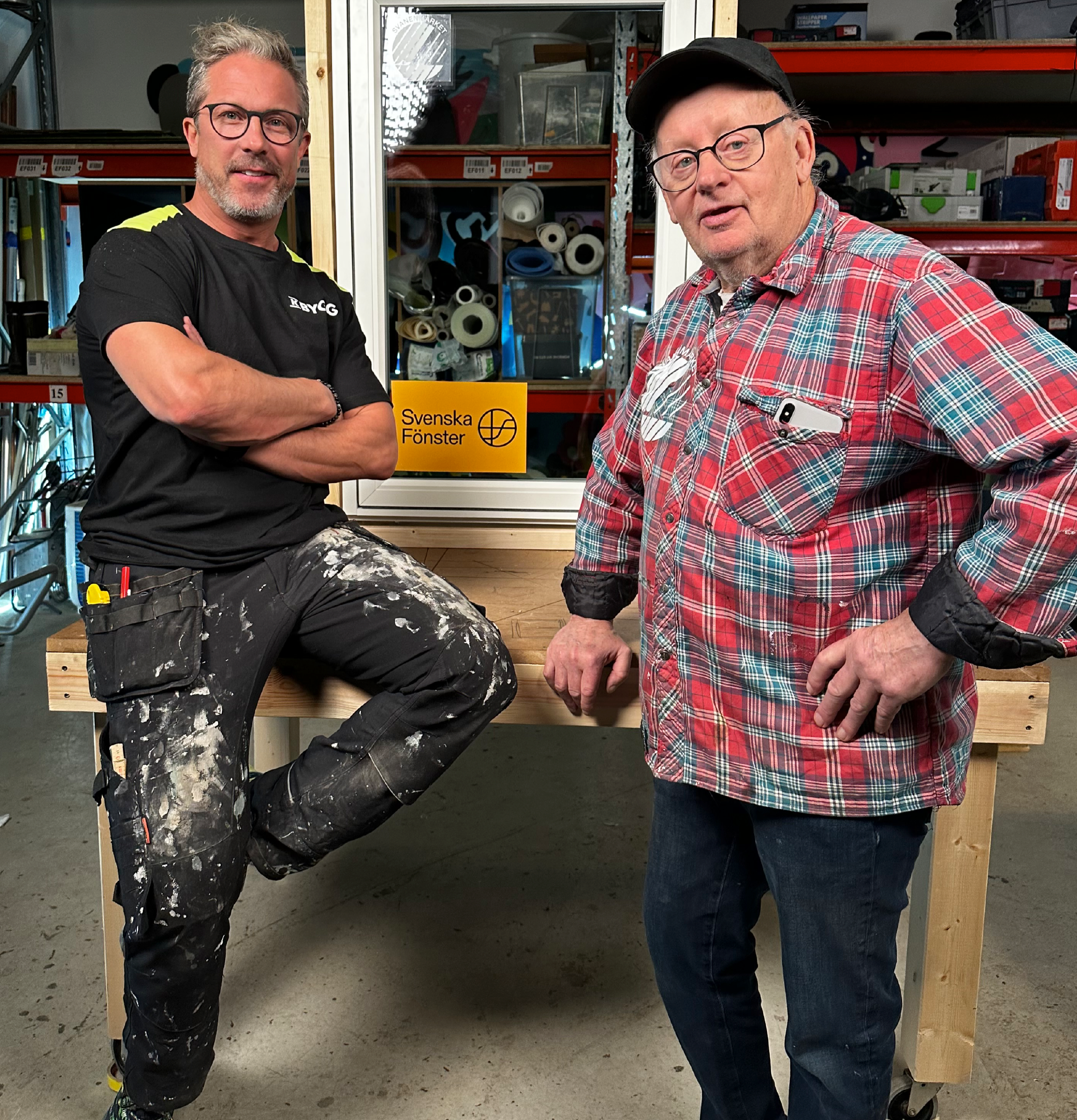 Två hantverkare står vid vitt fönster med "Svenska Fönster"-dekal i verkstad med röda hyllor.