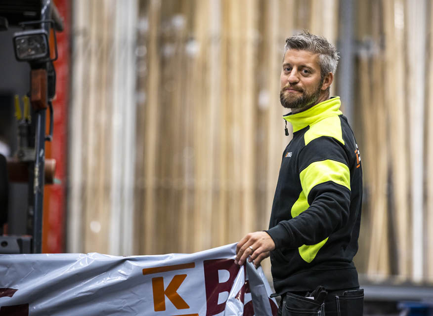 Byggarbetare i gul-svart jacka håller en plastduk med logotyp framför staplade träplankor.