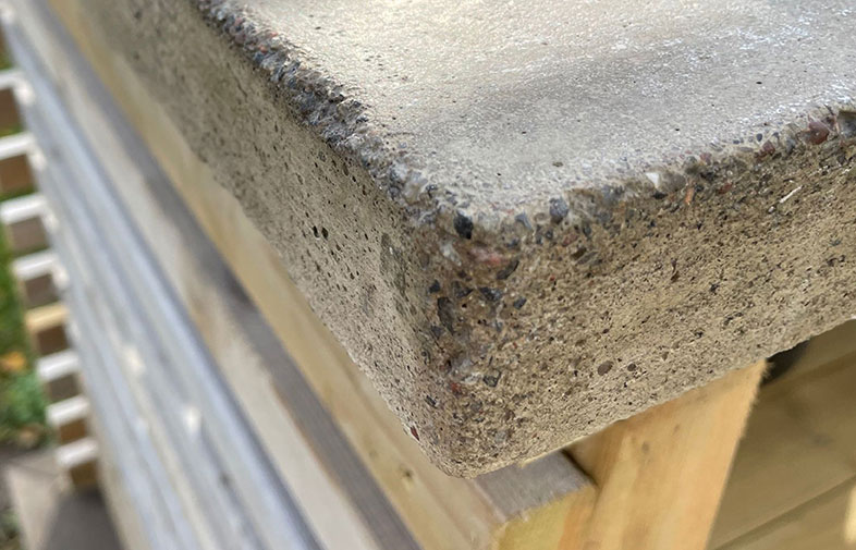 Närbild av grå betongkappa med synlig ballast på ljust trästaket.