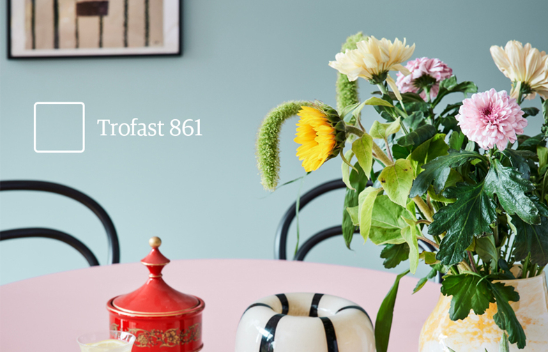 Runt rosa bord med bukett, rött krus, svarta stolar, turkos vägg och ’Trofast 861’.
