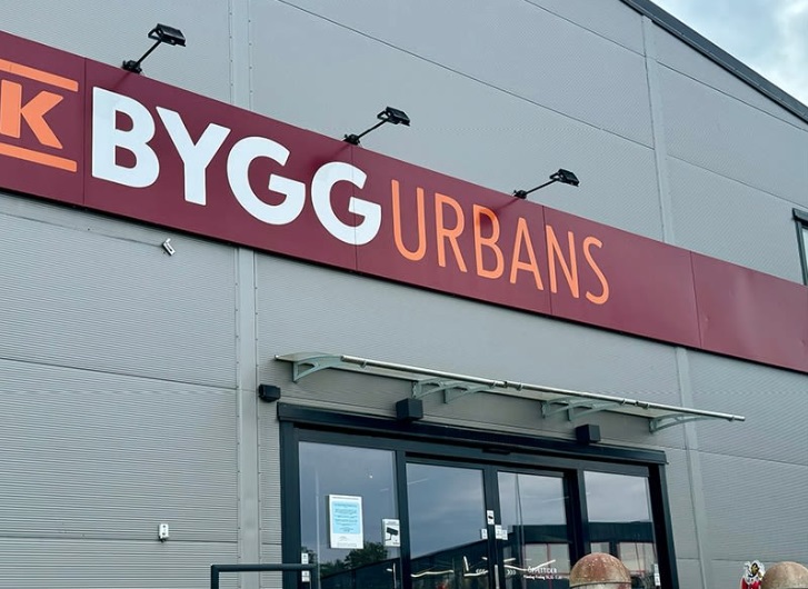 Fasad med rött skyltband ’BYGG Urbans’ i vitt och orange över glasdörrar på grå byggnad.