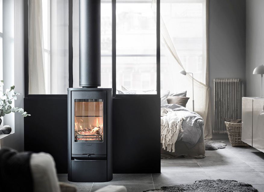 Svart rund kamin med sprakande eld framför glasvägg in mot lugn sovalkov med gråbeige textilier.