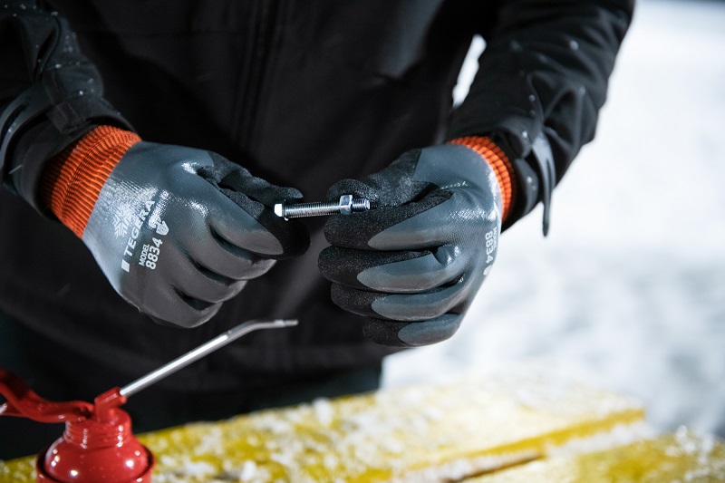 Svarta handskar med orange muddar håller en frostig metallbult över snötäckt bänk.