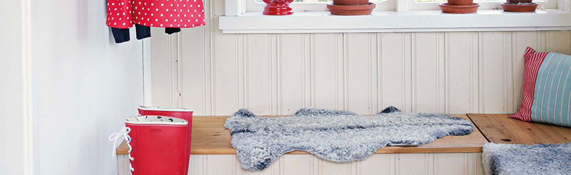 Inre entré: röd prickig regnkappa, röda gummistövlar, träbänk med grått fårskinn och randig kudde