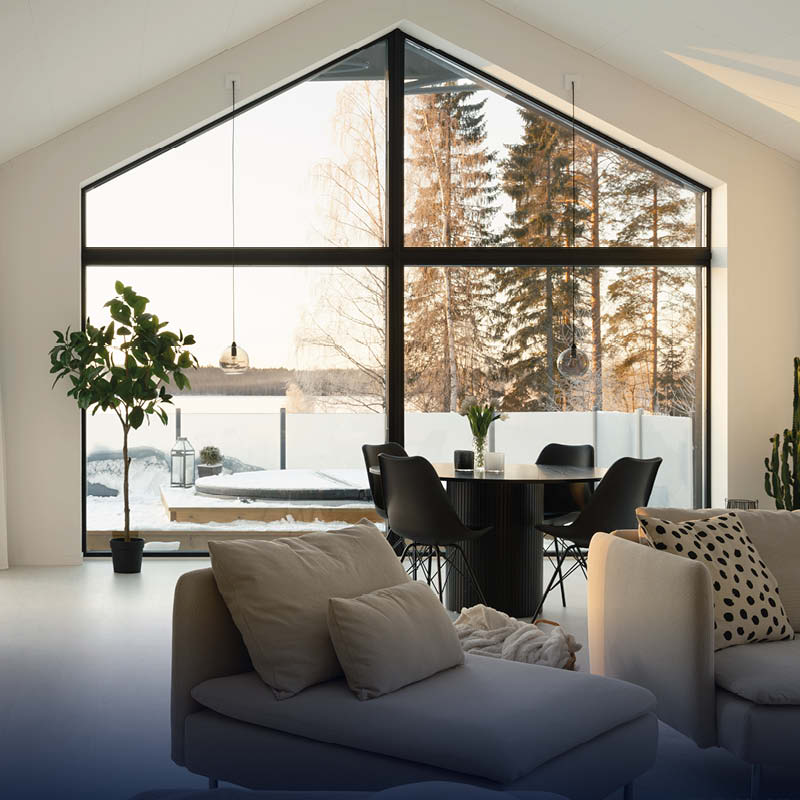 Ljust vardagsrum med stora triangelfönster mot snöig skog, beige soffa, svart matbord och växt.