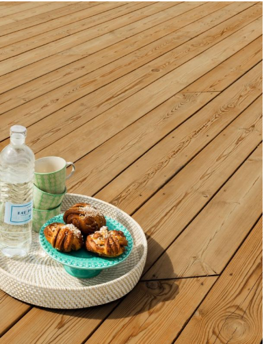 Vit flätad bricka på solbelyst trädäck med vattenflaska, grön mugg och kanelbullar på turkos tallrik