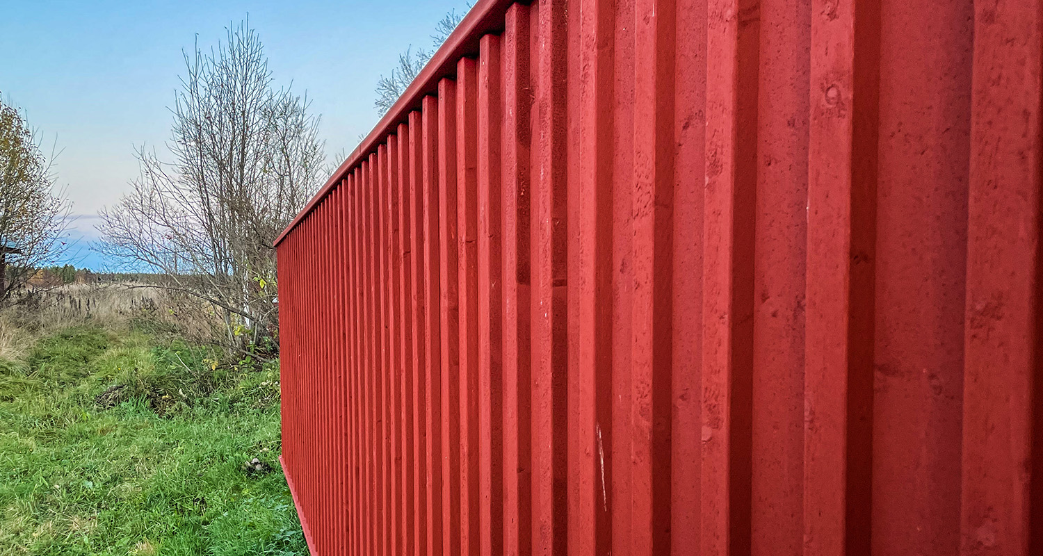 Röd korrugerad plåtvägg vid grönt gräs och kala buskar under klarblå himmel