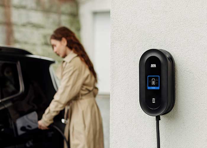 Svart elbilsladdare med blå display på vit vägg, kvinna i beige rock vid svart bil.