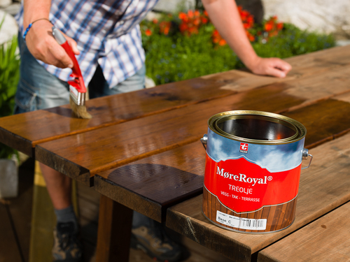 Person oljar mörkbrunt trädgårdsbord med pensel, och röd-blå MøreRoyal-oljeburk syns.