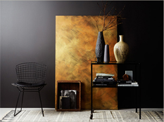 Svart trådstol, guldfärgad panel, svarta vaser med grenar, bokbord och beige vas mot grå vägg.