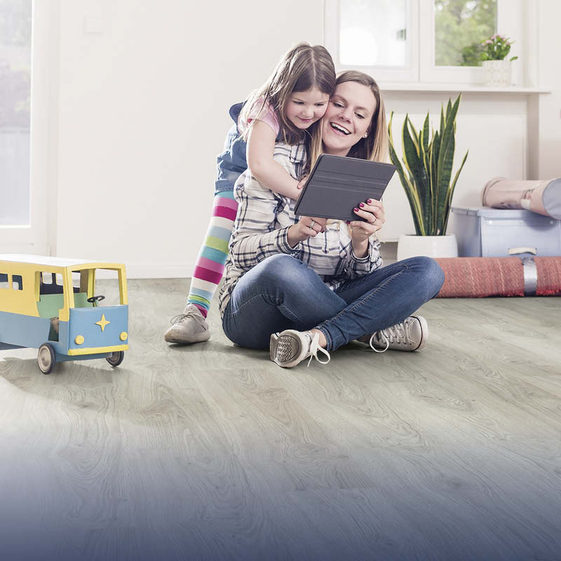Kvinna i rutig skjorta och jeans på ljust trägolv, barn i randiga byxor tittar på surfplatta.