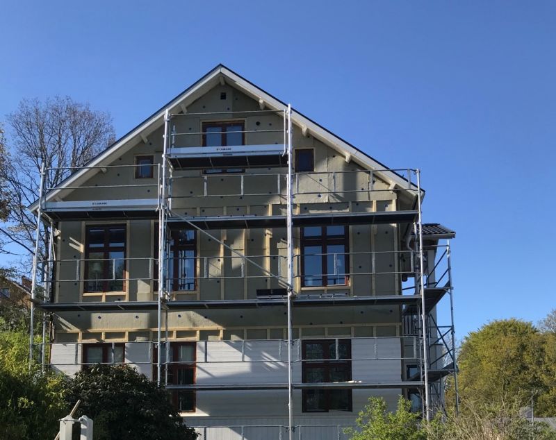 Trevåningshus under byggnadsställning med beige fasad, bruna fönster och klarblå himmel