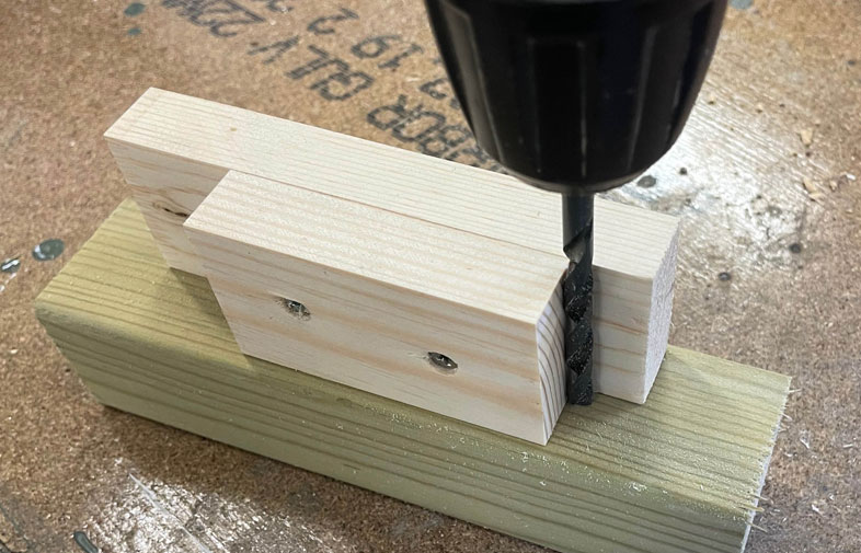 Svart spiralborr borrar i ljus träjigg på grönt träblock på dammig verkstadsbänk.