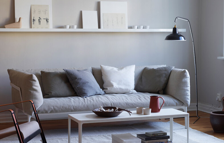 Ljust vardagsrum med grå soffa, vita och mörkgrå kuddar, vitt soffbord med röd kanna, svart golvlampa och konsthylla.