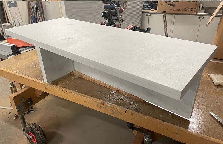 Låg gråmålad hyllsektion i trä på snickarbänk med verktyg och kartonger i bakgrunden.