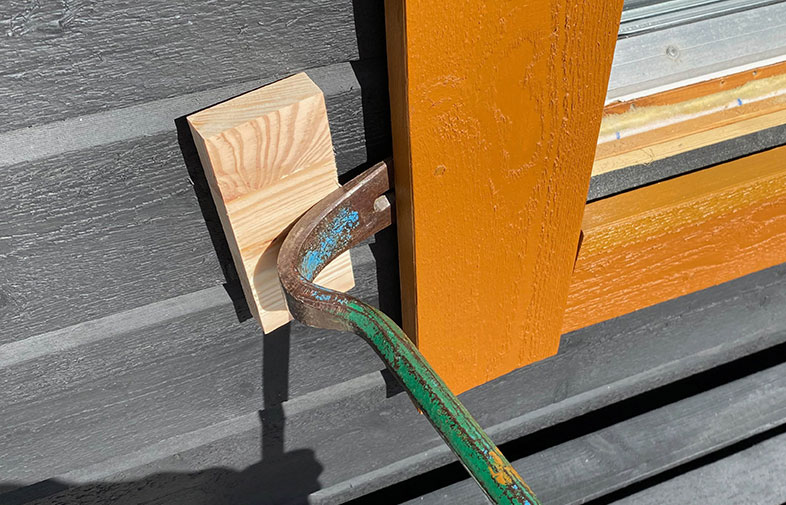 Grå vägg med orange fönsterkarm, ljus träkloss och en grön rostig kofot.