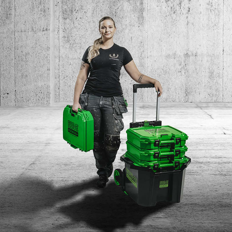 Kvinna i svart arbetsklädsel bär grön verktygslåda och rullar grönsvarta lådor på vagn.